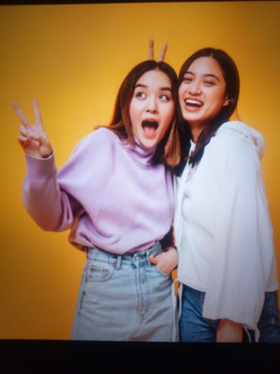 Two young women are posing for a photo against a solid yellow background. The woman on the left is wearing a light purple turtleneck sweater and light blue jeans. She has her mouth open and is holding up two fingers in a peace sign with her left hand. The woman on the right is wearing a white hoodie and blue jeans. She is smiling widely and has her hands on her hips, with one hand partially obscured by her hoodie. There are no discernible urban elements in the image that would indicate a specific location within посёлок опытного хозяйства «Ермолино», Russia.