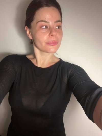 A woman with brown hair and eyes is posing. She is wearing a long-sleeved black top. Her skin appears dewy and the lighting enhances the texture. The image is taken from an upward angle, positioning her face in the upper left portion of the frame. The background is a plain white wall. Part of a tattoo is visible on her upper left arm.
