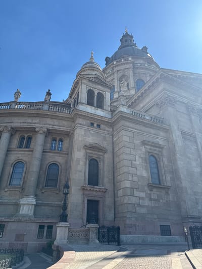 This is a low-angle, vertical photograph of the exterior of a large, ornate stone building, likely a cathedral or basilica, under a clear blue sky. The architecture features classical elements such as columns, arched windows, and elaborate carvings. A large dome with a spire tops the building, visible in the upper left portion of the image. The stone facade is detailed, with various window shapes and decorative cornices.
In the foreground, the paving stones of a plaza are visible, suggesting an outdoor public space. A wrought-iron fence or railing runs along the edge of the building, adding to the architectural details. Several small, statue-like figures are perched on ledges of the building.
The sun is bright, casting strong shadows, indicating it is likely daytime, possibly late morning or afternoon, given the angle of the light. The sky is a vibrant, cloudless blue, suggesting fair weather. The image's perspective looking upwards emphasizes the grandeur and height of the structure. There is no visible text in the image. The scene conveys a sense of architectural majesty and historical significance, possibly in a European city like Budapest, Hungary, as indicated by the location context.