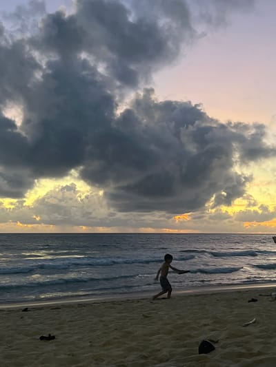 A boy is playing on a sandy beach with the ocean in the background. The sky is filled with dramatic, dark clouds that are lit from behind by the setting sun, creating a yellow and orange glow. The boy is running and appears to be holding a frisbee or a similar object. There are some pieces of driftwood and debris scattered on the sand. The water is dark and shows waves rolling towards the shore. The scene suggests a late afternoon or early evening setting. The location is not specified in the image or the prompt beyond a general geographic area.