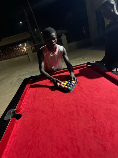 Two people play pool outside at night. One man in a white tank top holds a rack of pool balls, arranging them on the red felt of the pool table. Another person is above the table, partially obscured. The background shows a building lit by dim lights. The pool table is prominently featured, with the balls in focus. The lighting is low, creating shadows and highlights on the subjects.