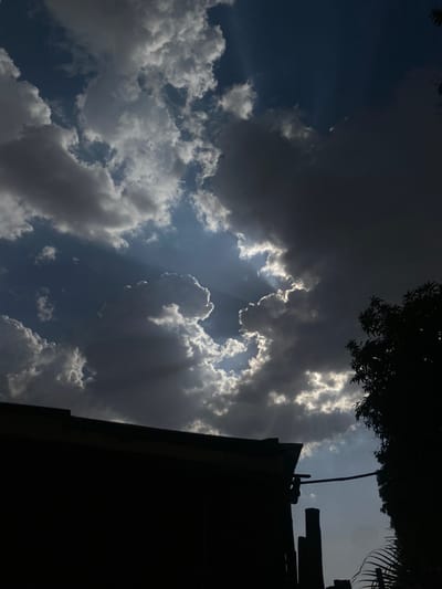The image, potentially taken in Barden Yana, Nigeria, presents a sky filled with a mix of dark and light clouds, with some rays of sunlight piercing through. The lower portion of the frame includes silhouetted trees and a structure, possibly part of a building.