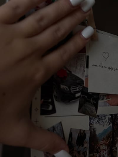 This media file features a close-up view of a hand with white fingernails, partially obscuring a collection of photographs. The photographs appear to be a mix of personal snapshots and possibly printed artwork. One visible photograph contains a hand-drawn heart and Cyrillic text that reads "от всего сердца," which translates to "from the whole heart." Another photograph shows a close-up of a black car. In the background, other photographs hint at outdoor scenes with trees and what looks like a garden or park. The overall lighting suggests it might be evening or indoors with low light. The presence of personal photographs and the "from the whole heart" message imply a sentimental context. The location context of Novosibirsk, Russia, is provided but not visually evident within the image itself.