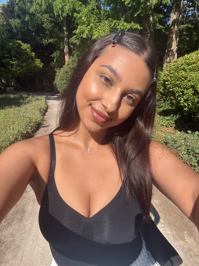 This is a selfie of a young woman in a park. She is wearing a black tank top and a silver cross necklace. Her hair is dark brown and pulled back with two silver clips. She is smiling and looking at the camera. The background is a park with green trees and a dirt path. There are flowers growing along the side of the path. The sun is shining, casting shadows through the trees. This park is located in Mönchengladbach, Germany.