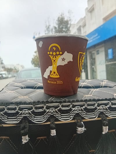 A close-up indoor shot shows a brown paper cup with a graphic of the continent of Africa, the outline of Morocco in white, and a golden trophy, labeled "Africa Cup of Nations Morocco 2025," resting on a black leather car seat cover with white stitching. The cover has a decorative braided trim with tassel-like accents. Outside the car window, a cityscape appears slightly out of focus under an overcast sky, featuring buildings and trees; judging by the cup details, the location is Martil, Morocco.