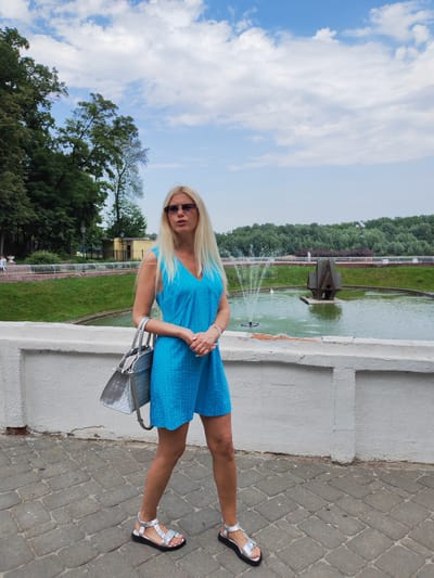 A woman with long blonde hair and sunglasses stands on a cobblestone path, facing the camera. She is wearing a short, bright blue dress with a V-neck and a subtle dotted pattern, and silver sandals. A large, metallic silver tote bag hangs from her left arm. Her hands are clasped in front of her.

Behind her, a white stone wall separates the path from a grassy area with a fountain in a pond. The fountain shoots water upwards. A decorative structure, resembling a small, dark, stylized house or building, sits in the center of the pond. In the background, a line of trees forms a dense green backdrop under a cloudy blue sky. The time of day appears to be daytime, with bright lighting suggesting fair weather. The setting is an outdoor public space, possibly a park or waterfront area, with elements of landscaping and water features. The woman's expression is neutral, and she appears to be posing for the photograph. No visible text is present in the image. The location context provided is Homyel, Belarus.