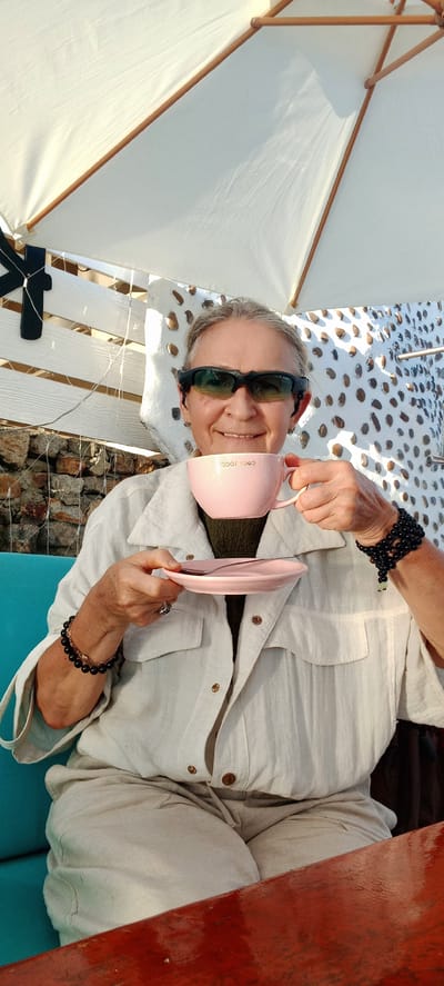The media file shows a person reclining on a white, slatted surface, possibly a bench or lounge chair, outdoors. A white umbrella provides shade. The person is wearing sunglasses and a light-colored, button-down shirt. They are holding a pink mug in their right hand and a pink saucer in their left hand, appearing to be in the process of drinking or having a beverage. The person is smiling, suggesting a relaxed or content mood. The setting appears to be a sunny outdoor location, likely during the day, with a glimpse of rocky ground and possibly a body of water in the background. Text on the mug reads "cocorococo". The overall impression is one of leisure and enjoyment in a warm climate.