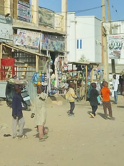 A dusty street scene showcases several young boys walking. One boy balances items on his head. The surrounding area is densely packed with small shops, displaying various goods and advertisements. A white building with two blue windows stands in the background. Utility poles are also visible. The overall atmosphere suggests a bustling, lively street in a developing area.