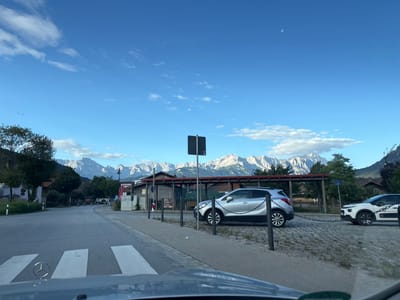The image shows a view from inside a vehicle looking out at a street in Farchant, Germany. A pedestrian crossing is visible in the foreground with white stripes on asphalt. To the right, there is a cobblestone area with two cars parked. One is a silver SUV, and the other is a white hatchback. Both cars are connected by a metal chain. Behind the cars, there are wooden structures and buildings, and a prominent row of snow-capped mountains forms the backdrop under a blue sky with some clouds and a crescent moon. Trees are present on the left side of the road and in front of the buildings. A street sign and a black pole with a sign are also visible. The overall scene appears to be a street view on a clear day with a mountainous landscape.