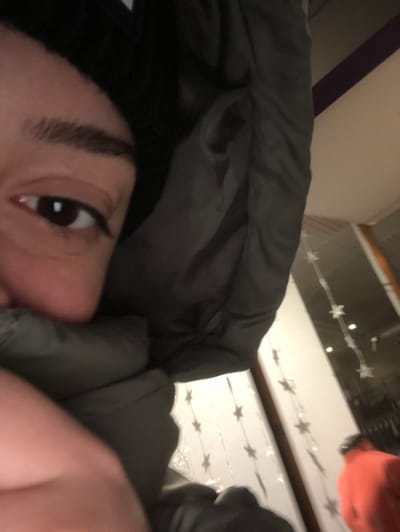 An extreme close-up shows a person's face partially obscured by a dark gray, puffy garment. The eye is visible, and brow appear well-defined. The background is blurry, but appears to be indoor setting with white horizontal surfaces. There are hanging decorations with star shapes visible. The orientation of the image is unusual, suggesting it was taken at an angle. Another person with orange shirt can be seen in the top right corner.