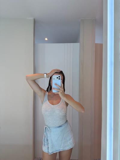 A person is taking a selfie in an interior space, which is presumably located in Ungasan, Indonesia. The person is wearing a light-colored tank top and blue denim shorts. They are holding a light blue smartphone up to their face, obscuring part of their features. Their left hand is holding their hair up behind their phone, and their right hand is covering their face. A white smartwatch is visible on their left wrist.
The background features interior architectural elements, including white walls, a ceiling with recessed lighting, and wooden beams. The lighting appears to be a combination of natural and artificial sources. A small, round light fixture is visible on the wall in the background. The space appears to be a well-lit, modern interior.