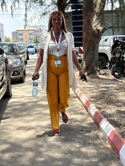 A woman stands outdoors near a street in Alaguntan, Nigeria. She holds a bottle of water in her left hand and a phone in her right. The woman has straight brown hair. She wears a white shirt, white jacket, and mustard-colored pants. Cars are parked on the street, and a building is visible in the background. There is a striped curb on the street. A motorcycle is parked near a tree trunk.