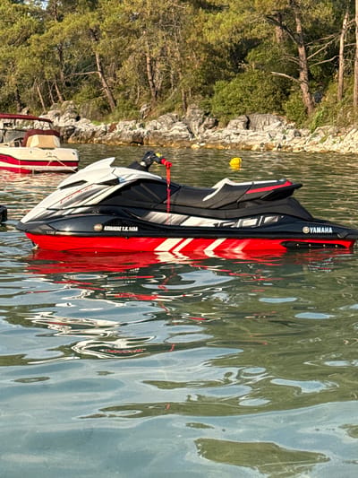 A jet ski, primarily black and red with white accents, floats on calm water. The Yamaha logo is visible on the side of the jet ski. In the background, a small boat with a red canopy is docked. The shoreline features rocky terrain and lush green trees. The scene appears to be a coastal area in the city of Thassos, Greece. There are no people visible in the image.