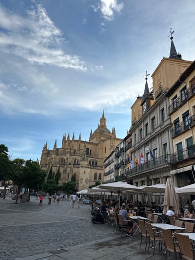 A partly cloudy sky overlooks buildings and a cathedral. One building has a clock and various flags. Outdoor dining with umbrellas and people occupy the right side of the image along the walkway. The cathedral is ornate with spires and windows. People walk along the cobblestone street at the base of the cathedral. Trees and greenery are visible at the bottom.