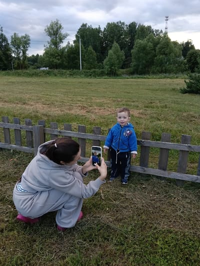 In a rural outdoor setting, a young child stands next to a wooden fence in a grassy field. The child is wearing a blue hooded jacket with a graphic print and patterned pants. A woman is crouched down in front of the fence, holding a smartphone and taking a picture of the child. The phone screen is visible, showing the child's image.

The scene appears to be in a natural environment with trees in the background and a cloudy sky overhead, suggesting it might be late afternoon or early evening. The grass is a mix of green and dry, with some patches of hay. A power pole or communication tower is visible in the distance to the right. The overall atmosphere seems calm and familial, with the woman focused on capturing a moment of her child. The location is identified as Smolino, Russia. No visible text is present in the image.