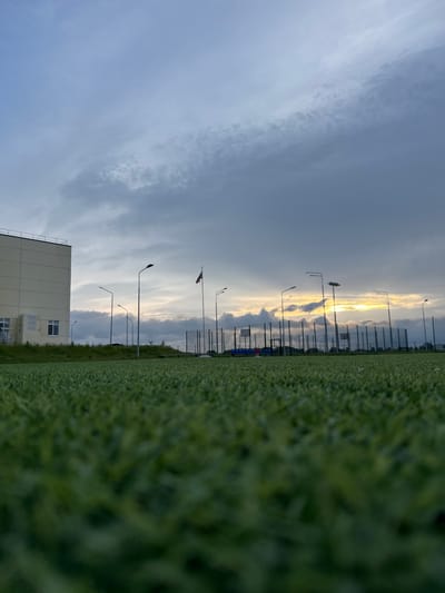 The image captures an outdoor scene in Ферма, Russia, featuring a low-angle perspective of an artificial green turf field that occupies the immediate foreground, gradually sharpening towards the midground. On the left side of the midground, a light-colored, multi-story building with multiple windows and external air conditioning units is visible. Extending across the midground from left to right is a series of tall, dark streetlights or floodlight poles. A tall flagpole displaying a red and white flag, indicative of the Russian national flag, stands between the building and the right side of the frame. Further to the right, a fenced outdoor sports court with a visible basketball backboard is present. Sparse green vegetation grows on a slight elevation behind the infrastructure. The sky in the background is largely overcast with grey and white clouds. A prominent yellow and orange glow, partially obscured by clouds, is visible on the horizon, consistent with either sunrise or sunset. No individuals are depicted in the scene.