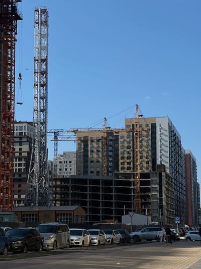 The image shows a construction site in Svkh Voronezhskiy, Russia. Several high-rise buildings are in various stages of completion, some with facades and others still under construction. Multiple cranes are present, including tower cranes and other construction equipment. A row of parked cars lines the street adjacent to the construction site. The sky is clear and blue. There is one bird in the sky. Several people are walking on the sidewalk.