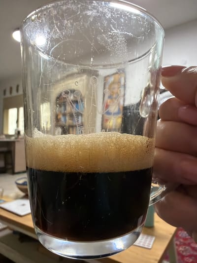 A hand holds a partially consumed dark beer in a glass mug, creating a distinct ring of creamy foam. The mug's surface is etched with visible scratches and an image of space ships. A reflection inside the glass shows a room interior. The beer itself is a deep, opaque black color. A small portion of a wooden table and patterned rug are visible in the background.