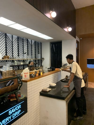 The image shows a cafe interior in Malang, Indonesia, located at -7.939 latitude and 112.631 longitude. A barista wearing a white shirt and black apron stands behind the counter, preparing a drink for a customer who is standing off-camera, holding a cup. The counter is made of black granite and white tiled wall, and behind it are shelves stocked with glass containers of coffee beans and other supplies. The barista is leaning forward, interacting with the customer. The cafe has a modern, minimalist design, with white walls, a black and white geometric patterned wall, and warm wood paneling. The ceiling is white and has recessed lighting. The cafe is clean and well-lit. The focus of the image is on the barista and the interaction with the customer. A sign on the left reads "SAVOR VERY DRIP". The sign on the right reads "FREE WiFi". There are also a few other objects in the image, including a coffee grinder, a shelf with glass containers, and a coffee machine. The cafe seems to be a popular local spot with both the barista and the customer present.