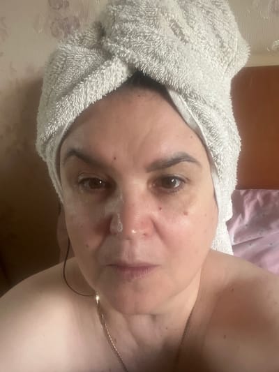 The image shows a close-up, rotated shot of a woman with a towel wrapped around her head. She has dark eyebrows and is wearing a subtle, shimmery eye shadow. Her skin appears to have some light blemishes and a bit of product on her face. She is wearing a delicate gold necklace. The background is blurred, but appears to be a domestic setting with soft lighting, possibly suggesting an evening or early morning. There are hints of fabric in pink and beige tones in the background, as well as a textured wall. The overall mood is relaxed and intimate, implying a personal moment of self-care. No text is visible in the image.