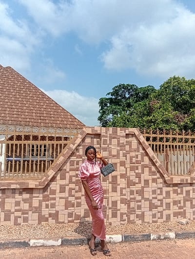 A young adult female is depicted standing outdoors in Ejule, Nigeria, posing in front of a tiled wall and metal fence. She wears a high-neck, long-sleeved, ruched pink satin dress and black open-toe flat sandals. Her left hand holds a dark, textured rectangular handbag, while her right hand is raised to the side of her head. The background features a low wall constructed from patterned light brown and beige tiles, topped by a decorative metal fence with intricate scrollwork. To the left, a building with a steep red-tiled roof is visible behind the fence. To the right, dense green foliage from trees extends above the fence line. The sky is light blue with scattered white clouds. The subject stands on a paved surface next to a curb, with gravel visible between the curb and the tiled wall.