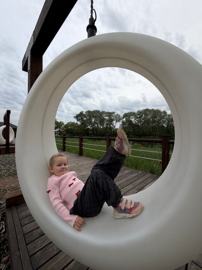 A young child is lying inside a white, circular playground structure in Kamyshin, Russia. The child wears a light pink sweater with small red bow accents, and dark pants with heart-shaped patterns. The child's feet, clad in worn sneakers, extend towards a background featuring a wooden deck, a green grassy area bordered by a low fence, and trees against a cloudy sky. The playground structure is supported by wooden beams. The ground around the structure is covered with wood chips and leaves.
