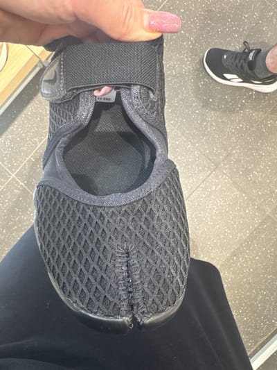 A person's hand, with light pink fingernails, is holding a black split-toe athletic shoe. The shoe is made of a mesh material with a diamond pattern. A price tag that reads "66.99" is visible on the shoe's strap. In the background, another black athletic shoe with white accents is on a person's foot, and a light-colored floor with speckled gray and white tiles is visible. There is no indication of the shoe's location in Poznan, Poland.