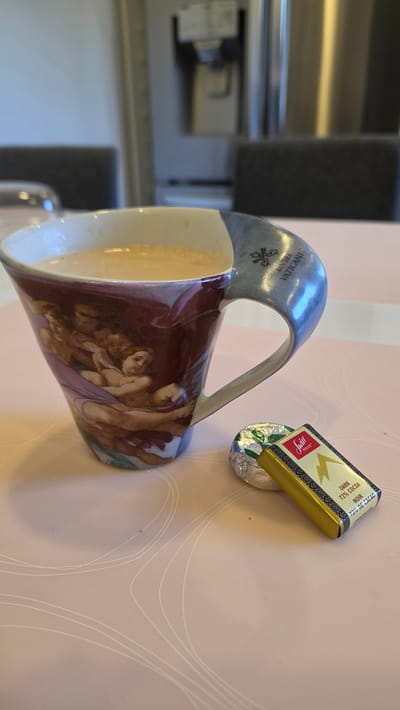 A detailed, forensic, matter-of-fact description of the scene:
The image shows a close-up, eye-level shot of a coffee cup and a small chocolate bar resting on a pink, patterned tablecloth. The coffee cup is ornately decorated with a depiction of cherubs in a classical art style, possibly referencing Michelangelo's Sistine Chapel ceiling, and features text that reads "MUSEI VATICANI" on its handle. The coffee within the cup is a light brown, suggesting it is coffee with milk or cream. To the right of the cup lies a miniature dark chocolate bar, still partially wrapped in gold foil, with the brand "Swiss Delice" and "DARK 72% COCOA" clearly visible on its packaging. The background is blurred, showing indistinct shapes of kitchen appliances and furniture, hinting at an indoor setting, likely a home or office. There are no people visible in the image. The lighting suggests an ambient indoor environment.
The location in the city of Toronto, Canada, cannot be determined from the provided image.