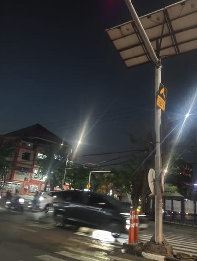 The image captures a nighttime street scene in Surabaya, Indonesia. A traffic intersection is visible, with a road marked by a crosswalk. Several vehicles, including cars and motorcycles, are in motion, their headlights and taillights creating streaks of light due to the long exposure.
In the foreground, a utility pole stands tall, equipped with solar panels at its top. A prominent yellow sign with a CCTV icon and text in Indonesian, "AREA DIAWASI CCTV," indicating a surveillance zone, is attached to the pole. Two orange traffic cones are placed near the base of the pole.
In the background, a multi-story building with red and white accents is visible, likely a commercial or institutional building. Trees and streetlights add to the urban ambiance. The sky is dark, suggesting it is late evening or early morning. The overall impression is one of a busy urban environment at night, with the presence of surveillance technology.