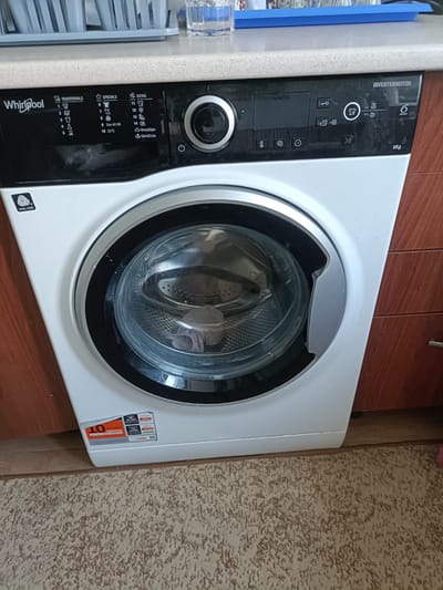 A front-loading Whirlpool washing machine is shown in a home setting, likely a kitchen or laundry room. The machine is white with a black control panel and a silver door. The control panel displays various wash cycle options labeled "TRADITIONALS," "SPECIALS," and "EXTRA." Symbols indicate different fabric types and wash settings, such as "WOOL CARE," "Eco 40-60," and "Rinse&Spin." The "INVERTERMOTOR" technology is also highlighted. To the left of the door, a sticker indicates "10 YEAR SERVICE WITH FREE PARTS." Below the control panel, there are additional icons for temperature, spin speed, and timer functions. The washing machine door is slightly ajar, revealing the drum inside. The machine sits on a light-colored tiled floor, with a patterned rug in the foreground. The background includes wooden cabinetry and a countertop with some kitchen items, suggesting an indoor domestic environment. No people are visible in the image. The lighting appears to be indoor artificial light.