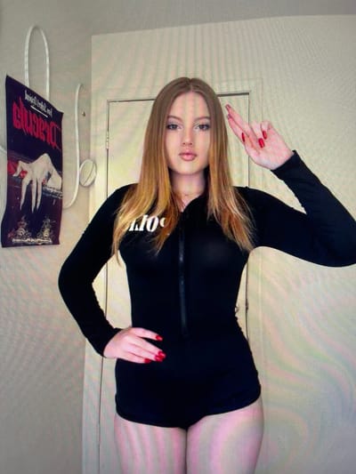 A young woman with long, reddish-blonde hair stands in front of a door, her left hand on her hip and her right hand raised with two fingers crossed. She is wearing a black, long-sleeved, zippered bodysuit. A poster with a dark background and a red, stylized title is visible on the wall to her left. The environment appears to be an indoor residential space. There is no information available to determine the specific location within the city of Łódź, Poland.