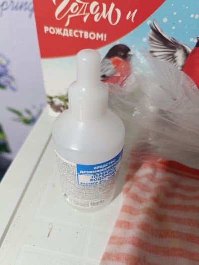 A white plastic bottle of hydrogen peroxide solution is on a white surface, next to a red and white striped fabric. In the background, a postcard or printed material depicts birds and holiday greetings. The overall scene appears to be indoors, possibly in a domestic setting. The image does not provide any specific location information within the city of Nikolskoye, Russia.