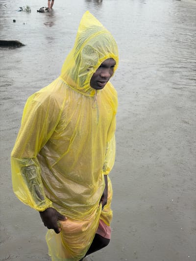 The image shows a person wearing a bright yellow, translucent plastic raincoat with a hood, standing in shallow, murky water during a rainstorm. The person is looking down and to their right, with their left hand inside the raincoat, touching their right thigh. Their face is visible within the hood. The raincoat is covered in water droplets, indicating the rain is active. The environment appears to be an outdoor urban area in Igboefon, Nigeria, with the ground flooded with rainwater. In the background, other figures and debris are visible in the water. The overall scene depicts a rainy day with significant flooding.