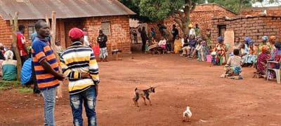 In a rural setting with red soil, a gathering of people is observed. Most of the attendees are seated in a circle, with brick buildings in the background. In the foreground, two individuals are standing, facing left. A goat and a duck are also visible in the open space, adding to the rural atmosphere. The scene appears to be a community event or meeting.