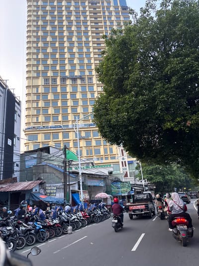 The image depicts a street scene in Central Jakarta, Indonesia. In the background, a tall, modern building with a repetitive facade of light-colored panels and blue windows dominates the skyline. In the foreground, a row of parked motorcycles lines the side of the road. Several people are visible, some on motorcycles and others standing near the parked vehicles. A black pickup truck with "LALAMOVE" branding is in motion on the street, as are other motorcycles and cars. Trees with dense green foliage are present, casting shadows on the street. Various signs and awnings are visible on the buildings along the roadside, suggesting a commercial or residential area. The overall scene captures a bustling urban environment with traffic and pedestrian activity.