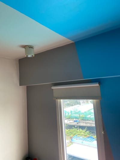 The image shows an interior view of a room with a distinct color-blocked design on the walls and ceiling. The ceiling is split diagonally, with the upper portion painted bright blue and the lower portion a lighter shade. A white cylindrical light fixture is mounted on the lighter section of the ceiling. The walls are also painted in contrasting colors: a light cream color on the left, a medium gray in the center, and a bright blue on the right. A window with a roller blind is visible in the center right of the image. The view outside the window shows buildings, greenery, and what appear to be greenhouses or construction sites, suggesting an urban environment. There are no people visible in the image. The lighting suggests it is daytime. Without further information, the specific location in Central Jakarta, Indonesia cannot be determined from the image alone.