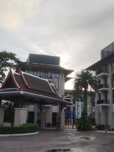 The image shows the entrance to a resort or hotel in Ko Kaeo, Thailand. The architecture features a prominent traditional Thai roof with red and white trim, supported by pillars. This entrance structure is in the foreground and to the left. Behind it, to the left and center, is a multi-story building with a dark, sloped roof. To the right and center, another multi-story building is visible with balconies and dark railings. Several palm trees are present, including one tall, slender palm in front of the building on the right, and a wider palm to the left of the same building. The ground in front of the entrance is paved with cobblestones, and some areas are wet, indicating recent rain. The sky is overcast with diffused light, suggesting it is either early morning or late afternoon. There are no people visible in the scene.