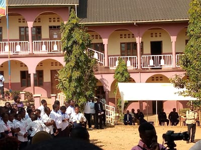 The image shows a large gathering of people in front of a building in Beni, Democratic Republic of Congo, located at the coordinates (latitude: 0.492, longitude: 29.483). The building is two-story with a pink facade and arched openings, with a white railing around the second floor. There are trees and green vegetation around the building. A white canopy tent is set up in front of the building. A group of people are standing in front of the building, some of whom are dressed in white shirts with badges, and others are in suits or casual clothing. There is a man in the foreground, holding a video camera, and filming the people in the gathering.