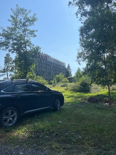 The image depicts an abandoned, multi-story building with a skeletal appearance, its concrete structure exposed and balconies open to the elements. This structure stands amidst lush greenery, with tall trees and dense bushes surrounding it. In the foreground, a dark-colored car is parked on a gravelly area adjacent to a grassy slope. The car is partially obscured by foliage, with its tires and lower body visible. The scene is bathed in bright sunlight, suggesting it is daytime. The sky is clear and blue. The overall impression is one of decay and nature reclaiming a man-made structure. The location context provided is Ureki, Georgia, which is known for its sandy beaches and Soviet-era architecture. No people are visible in the image. There is no discernible text in the image.