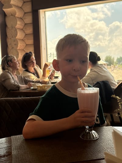 A young Caucasian boy with blond hair is seated at a table in a restaurant, holding a glass of pink milkshake and drinking it with a straw. He is wearing a dark green t-shirt with white trim. In the background, two women are seated at the same table, and another person with dark hair is visible further back. The restaurant interior features a wooden wall and a large window looking out onto a cloudy sky and a landscape that appears to be a farm. The lighting suggests it is daytime. The overall setting is casual and appears to be a family outing.