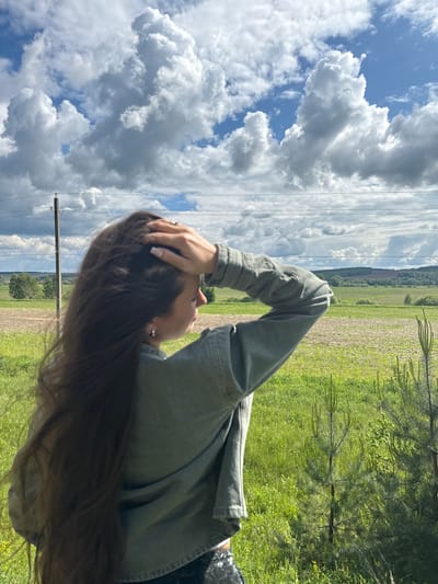 A medium shot depicts a woman outdoors, likely in a rural setting near Kremyonki, Russia. She is turned toward the left, head tilted up, with her left hand resting on her head. She wears a long-sleeved, light green jacket. Her hair is long and brown. The sky is cloudy with patches of blue. The landscape consists of green fields and some distant trees. Utility poles and lines are visible. A few small pine trees are in the foreground.
