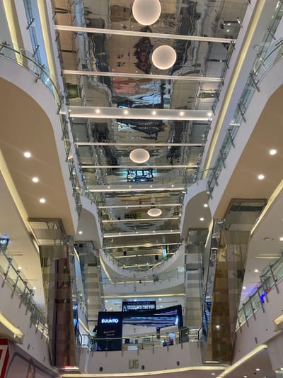 The image depicts the interior of a multi-story shopping mall, likely located in West Jakarta, Indonesia, given the context provided. The perspective is looking upwards, showcasing the architectural design of the mall. The ceiling is a highly reflective, metallic surface with a corrugated or ridged texture, creating a distorted, mirrored effect of the floors and elements above. Several large, round, white light fixtures are visible on the ceiling.
The mall has multiple levels with curved walkways and balconies, all featuring glass balustrades supported by metal railings. The walls of the mall are predominantly white and cream-colored, with some sections clad in polished, reflective materials, possibly stainless steel or mirrored panels, which reflect the surrounding environment. Integrated lighting strips are present along the edges of the walkways and ceilings, contributing to the overall bright and modern ambiance.
Visible on the lower levels are store fronts and signage. One prominent sign displays "SUUNTO" with the tagline "Adventure starts here". Another sign reads "UG", possibly indicating a floor number or a mall designation. A few individuals are faintly visible on the different levels, suggesting the mall is open and operational, though they are not the primary focus of the image. The overall scene conveys a sense of spaciousness, modernity, and visual complexity due to the extensive use of reflective surfaces.