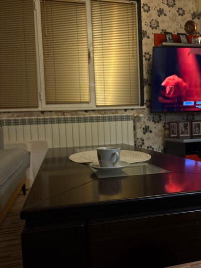 An interior residential scene in Cherven Bryag, Bulgaria, features a dim living area. A dark, reflective coffee table occupies the foreground, supporting a white ceramic cup containing dark liquid, resting on a matching saucer atop a circular woven placemat. A clear glass panel is integrated into the tabletop surface beneath the cup. To the left, the armrest and portion of a light-colored sofa are visible. The background reveals three windows fitted with closed, gold-toned horizontal Venetian blinds, positioned above a long white panel radiator and a rough-textured wall section. On the right, a wall covered in dark floral patterned wallpaper displays a wall-mounted flat-screen television showing an image with prominent red tones, depicting a hand or object, alongside minimal on-screen text. Above the television, a narrow reddish-orange shelf holds several small framed pictures, with a circular metallic decoration mounted higher on the wall. A dark console with additional framed items is partially visible below the television. No people are present, and no actions or interactions are depicted.