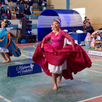 A woman in a red dress and white petticoat dances barefoot on a green floor with multicolored lines. To her right is a blue sign with white lettering that says "Marineras con Tradition Revista" with an illustration of a woman and man dancing. To the left, speakers sit in front of a draped blue and white backdrop, behind which, people are seated. In the background, more people are seated in the audience. Above the dancers is a banner that reads "MUNICIPALIDAD DISTRITAL DE MIRAFLORES."