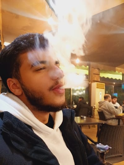 A man with a beard exhales smoke. He is wearing a black jacket over a white hooded sweatshirt. The background is a blurry mix of yellow, green, and brown hues. Other people sit in the background, indistinct. The scene seems to be indoors, possibly a cafe or lounge. The man's face is the focal point, partially obscured by the smoke he's releasing.