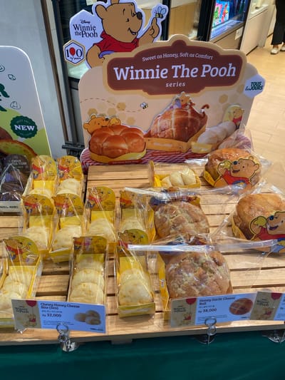 This image shows a display of baked goods, likely from a bakery, with a Winnie the Pooh theme. The products are arranged on wooden shelves and are individually wrapped in clear plastic. Many of the packages feature illustrations of Winnie the Pooh and friends.
The prominent display sign on the left reads "Disney Winnie The Pooh series," with smaller text "Sweet as Honey, Soft as Comfort." It also mentions "100 years," suggesting a celebration related to Winnie the Pooh's anniversary.
Several types of bread and pastries are visible. One sign indicates "Soft Honey Butter Bread." Other visible product names include "Honey Garlic Cheese Ball" priced at Rp 38,000, and "Chewy Honey Cheese Bite (3ea)" priced at Rp 32,000. The text on these signs includes both English and Korean.
The overall setting appears to be indoors, likely a retail environment. The lighting suggests it could be daytime. The presence of price tags in Indonesian Rupiah (Rp) and the text "North Jakarta, Indonesia" in the context clues confirm the location. There are no people clearly visible in the frame, focusing entirely on the food products and promotional materials. The scene is clean and organized, typical of a bakery display.