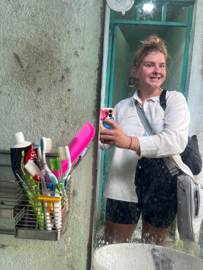 A young woman with blonde hair in a messy bun smiles as she takes a selfie in a mirror. She is wearing a white button-down shirt and black shorts. A crossbody bag is slung over her shoulder. In the foreground, a metal basket holds several toothbrushes and a tube of toothpaste. The background shows a green-framed doorway and a textured, off-white wall. The scene appears to be a bathroom in a budget hotel or guesthouse in Bo Y, Vietnam.