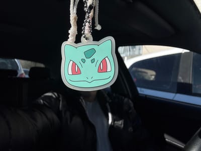 This image depicts the interior of a vehicle on a bright, sunny day in Rennes, France. A stylized, light teal-colored Bulbasaur head, functioning as an air freshener or charm, hangs centrally in the foreground, suspended by a white braided string and a silver ball chain. The Bulbasaur features prominent red eyes with white pupils and darker green spots on its head. A blurred individual, dressed in a dark puffer jacket over a lighter shirt, is visible in the driver's seat. Through the car's windows, other vehicles are observable in the exterior environment; a white car with tinted windows is prominent on the right, and another blurred vehicle is partially visible on the left. The sun illuminates the scene from the front and right side of the vehicle.