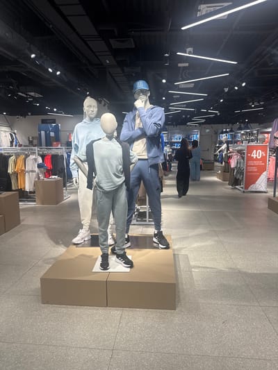 This media file depicts a retail store interior, likely a clothing store, with a focus on athletic wear. In the foreground, there are several mannequins dressed in casual sportswear. The mannequins are arranged in a way that suggests they are displaying outfits.
One mannequin is wearing a blue tracksuit and a blue cap. Next to it, another mannequin is dressed in a light blue hoodie and light-colored pants, paired with white sneakers. A third mannequin is wearing a light blue top and dark gray pants, also with black sneakers. The clothing displays are placed on neutral-colored pedestals.
In the background, racks of clothing are visible, with various t-shirts in different colors, including red, yellow, and orange. A prominent sign in the background features a "40%" discount offer, with text in what appears to be a Cyrillic-based alphabet, "2 НЕМЕСЕ ОДАН ДА КӨП ЗАТТАРДЫ САТЫП АЛҒАНДА ПРИ ПОКУПКЕ 2 И БОЛЕЕ ВЕЩЕЙ," which translates to "When buying 2 or more items." This indicates a sale or promotion.
The store appears to be well-lit with artificial lighting, including long strip lights on the ceiling and spotlights directed at the displays. The floor is tiled with a speckled gray pattern. The overall atmosphere suggests a modern and organized retail environment. The context provided indicates the location is Kokshetau, Kazakhstan. There are no discernible elements suggesting time of day or weather conditions, as the scene is indoors. The emotions conveyed are neutral, typical of a commercial setting.