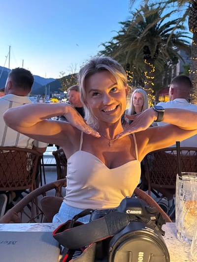 A blonde woman with fair skin and bright blue eyes is seated at an outdoor table at dusk in Tivat, Montenegro. She is smiling and looking at the camera, her left hand near her neck and her right hand resting on her chest. She is wearing a light-colored tank top with thin straps and a cross necklace. Her arms are visible, along with a black Apple Watch on her right wrist. In front of her, on the table, is a black Canon EOS camera with a strap.
In the background, other people are seated at tables. To the left, a man wearing a striped shirt is partially visible, and further left, a boat mast is seen against the twilight sky. To the right of the woman, a blonde woman with light skin and blue eyes is seated, looking ahead. In the far right of the background, another man in a white shirt is visible.
The setting appears to be a restaurant or bar with palm trees adorned with fairy lights in the background. The sky is a deep blue, indicating it is evening. The table has a marble-like surface. A glass with ice and a drink is on the right side of the table. The chairs are made of wicker. The overall atmosphere is social and relaxed.